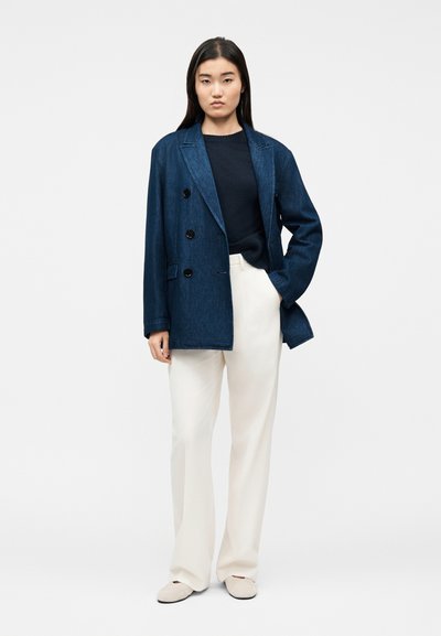 Γυναίκα που φοράει σκούρο μπλε σακάκι διπλής σειράς, μαύρο τοπ, λευκό παντελόνι με φαρδιά πόδια και λευκά παπούτσια, στέκεται με το ένα χέρι στην τσέπη.