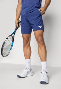 Tume merev murtide lühikesed püksid koos valge logoga, valged tennisekingad ja lühikesed sokid. Ühes käes hoitakse musta ja türkiissinise tennisereketit.