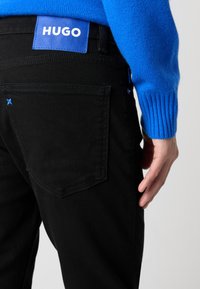 Los jeans de mezclilla negros presentan un parche con el logo azul de HUGO en la cintura, con detalles de costura y un acento azul en el bolsillo trasero.