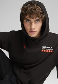 Sudadera negra de tejido suave, con un logo rojo de Puma y el texto "FORMULA 1 DRIVE TO SURVIVE" acompañado de cinco puntos rojos.