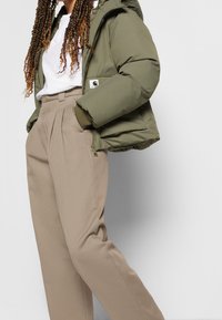 Persoon met lang gedraaid haar die een olijfgroene pufferjack, wit t-shirt en beige hoge geplooide broek draagt met de handen in de zakken.