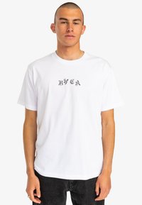 White cotton t-shirt with short sleeves, crew neck, and a black stylized logo printed on the chest. Smooth texture and relaxed fit.