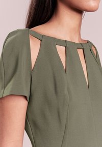 Oliwkowa sukienka z krótkimi rękawami i geometrycznym wycięciem w okolicy dekoltu, wykonana z gładkiego materiału i dopasowanego kroju.