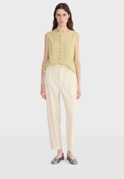 Femme portant un chemisier sans manches jaune à carreaux, un pantalon taille haute crème et des ballerines argentées métalliques, debout devant un fond uni.