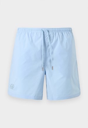 Jeune homme portant des lunettes de soleil noires, un débardeur blanc, un short bleu clair, des chaussettes blanches et des baskets écrues, debout les mains dans les poches.