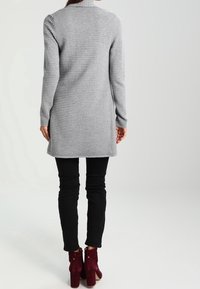Abito lungo grigio con maniche a manica lunga, texture a costine, colletto alto e taglio sopra il ginocchio, abbinato a pantaloni neri slim-fit e stivali alla caviglia bordeaux.