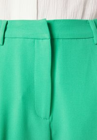 Close-up van hoog-taille turkooizen broek met een verborgen ritssluiting aan de voorkant en lusjes voor een riem, gecombineerd met een witte geribbelde top.