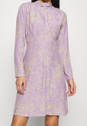 Kvinde iført en langærmet, højhalset kjole i knælængde i lilla med et lyst gult leopardmønster.