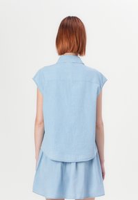 Camicia di jeans azzurra chiara con maniche corte, colletto classico e orlo arrotondato. Il tessuto appare morbido con una leggera texture.