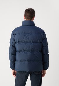 Marineblå pufferjakke med høj krave, quiltet tekstur og elastiske manchetter. Båret med mørke jeans, set fra ryggen.
