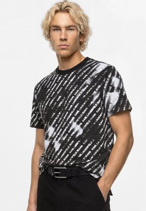 Junger Mann mit blondem lockigem Haar, der ein schwarz-weiß gemustertes Karl Lagerfeld T-Shirt und schwarze Hose mit Gürtel trägt.