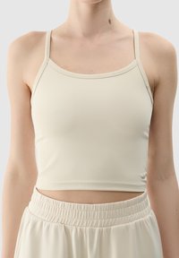 Top corto beige sin mangas con tirantes finos, hecho de una tela suave y elástica. El diseño presenta un escote redondeado y costuras mínimas.