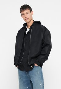 Chaqueta bomber negra con capucha, cierre de cremallera y puños acanalados. Llevada sobre una camisa de botones y combinada con jeans azul desgastados.