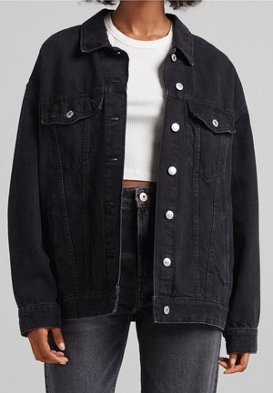 Person iført oversized sort denimjakke over hvid ribbet croppet top og sort high-waisted jeans mod grå baggrund.