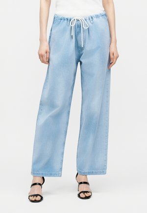 Άτομο που φοράει ανοιχτό μπλε τζιν παντελόνι με φαρδιά πόδια, λευκό κορδόνι στη μέση και μαύρα πέδιλα με ανοιχτά δάχτυλα και τακούνι.