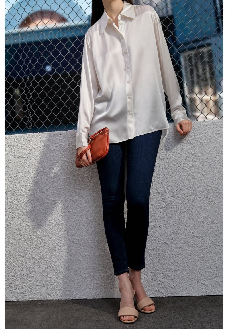 Camisa blanca de satén con botones, jeans oscuros ajustados, sandalias beige de slide y un pequeño bolso cruzado naranja, posando frente a una pared texturizada.