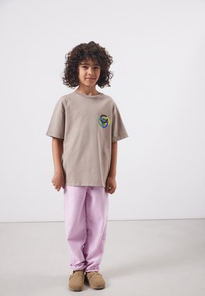Kind mit lockigem Haar, das ein beige übergroßes T-Shirt mit buntem Logo, rosa Cordhosen und braune Slipper trägt, steht drinnen.