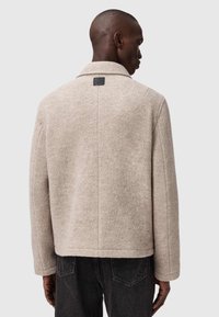 Beige uldblandingsjakke med notched krave, minimal syning og firkantet skulderdesign. Har et firkantet mærkelabel på ryggen.