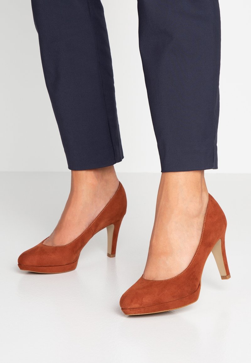 Femme portant un pantalon bleu marine et des chaussures à talons hauts en suède couleur rouille, debout sur un sol blanc.