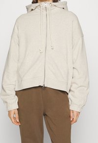 Felpa cropped beige in morbido tessuto, con zip frontale, cappuccio regolabile e polsini a costine. Sono visibili due tasche frontali.
