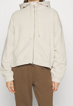 Felpa cropped beige in morbido tessuto, con zip frontale, cappuccio regolabile e polsini a costine. Sono visibili due tasche frontali.