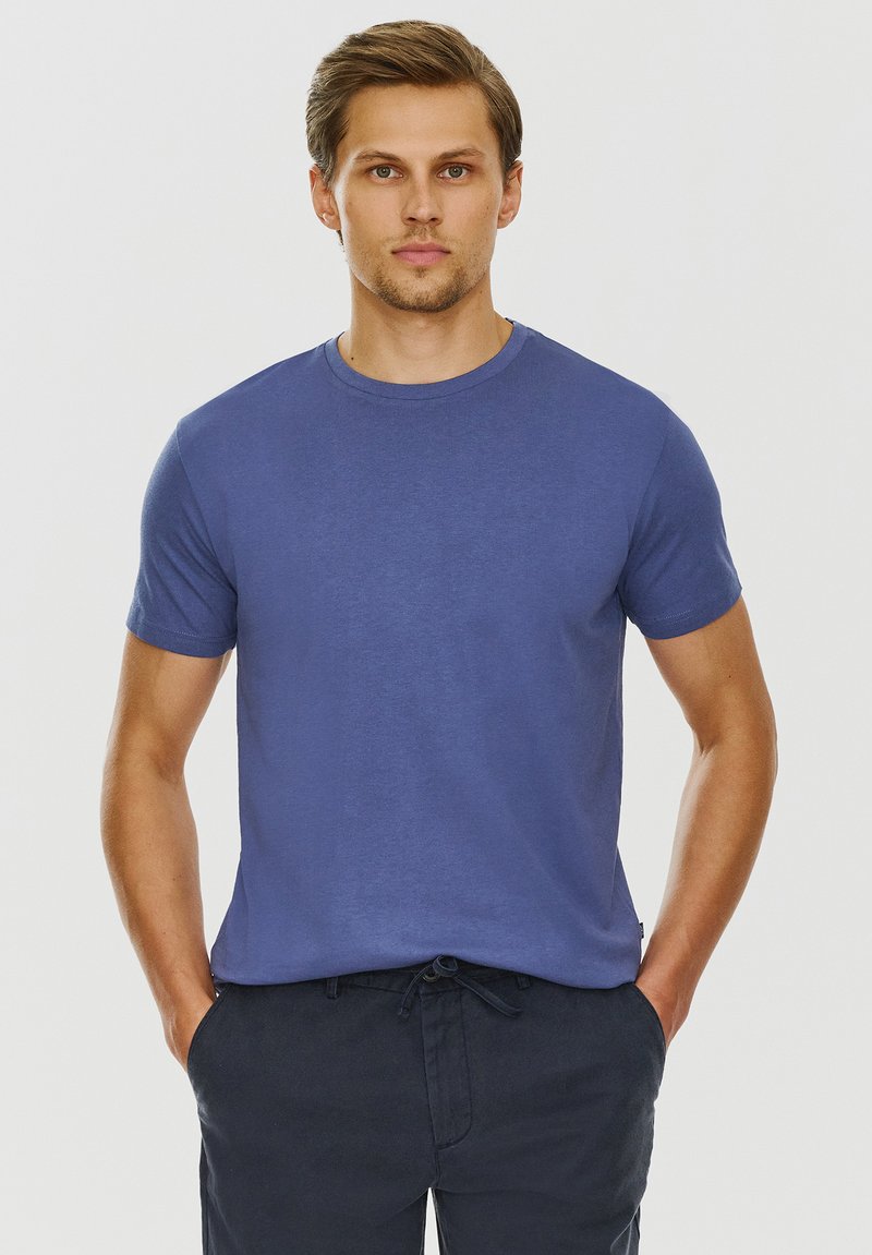 Niebieska bawełniana koszulka z okrągłym dekoltem i krótkimi rękawami, o gładkiej fakturze i luźnym kroju. Noszona z ciemnymi spodniami.