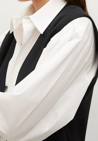 Schwarze ärmellose Weste über einem weißen Button-up-Hemd, das die kontrastierenden Farben und geschichteten Stoffe betont. Verfügt über weite Ärmel und einen spitzen Kragen.