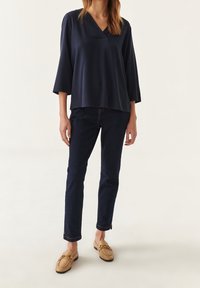 Haut bleu marine à col en V et manches longues, fabriqué dans un tissu lisse, associé à un jean en denim foncé et des mocassins beiges avec des accents en métal doré.