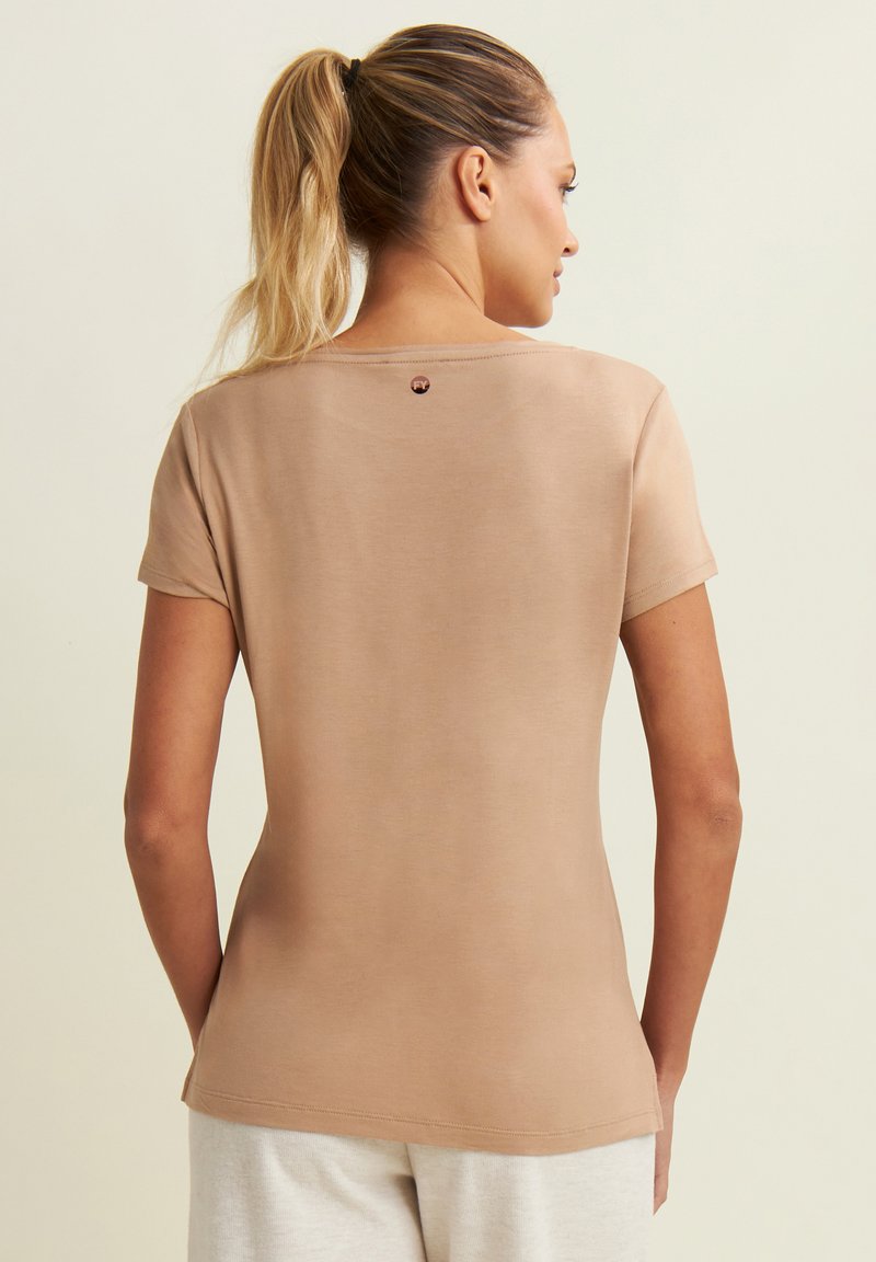 Camiseta de manga corta color beige con una textura suave, cuello redondo y un pequeño detalle del logo en la parte posterior. Silueta ajustada, diseño minimalista.