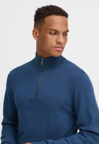 Mørkeblå zip-neck genser med ribbestrikket krage og strukturert strikket materiale. Har en halv-zip lukking og lange ermer.