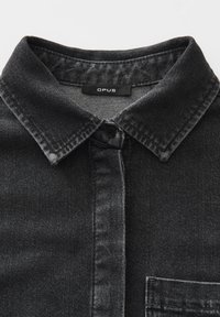Schwarze Denim-Hemd mit Stehkragen, oberem Knopf geschlossen und sichtbarer Brusttasche, im Halsausschnitt mit einem Etikett "OPUS".