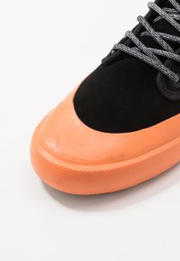 Schwarzer Sneaker mit glänzender, oranger Gummispitze, strukturierter Sohle und schwarzen Schnürsenkeln mit weißen Sprenkeln. Sichtbare Materialien sind Wildleder und Gummi.
