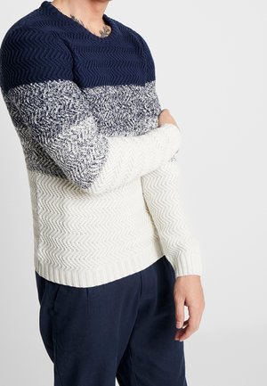 Mężczyzna ubrany w sweter z fakturowanym splotem z blokami kolorów granatowego, szarego i białego, w połączeniu z ciemnogranatowymi spodniami, na jednolitym białym tle.