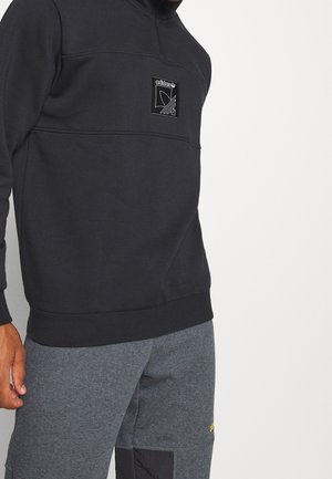 Homme portant un sweat-shirt Adidas noir avec un patch logo sur la poitrine et un pantalon de survêtement gris foncé, debout devant un fond blanc.