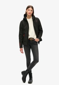 Zwarte softshell jas met rode accenten, ritszakken en een capuchon, gedragen over een crèmekleurige trui met een donkere spijkerbroek en zwarte veterschoenen.