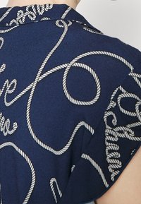 Marininspirerad skjorta i blått tyg med vitt repmönster och rundad krage. Baksidan visar flödande linjer och slingor av designen.