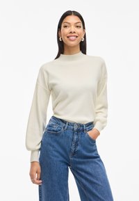 Cream-colored, high-neck sweater with loose, long sleeves, paired with high-waisted blue denim jeans. Soft texture, relaxed fit.