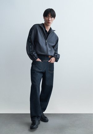 Ung mand stående med hænderne i lommerne iført en grå lynlåstrøje med sorte striber og mørke løse jeans med sorte sneakers.