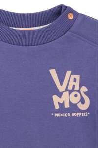 Lila sweatshirt med ribbat krage, med ljusbeige text som står "VAMOS" och "MEXICO NOPPIES." Knappliknande accent nära halslinningen.