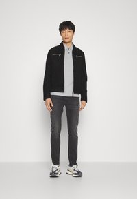 Chaqueta de ante negra con dos bolsillos con cremallera, camiseta polo gris, jeans gris oscuro y zapatillas navy con detalles en blanco. Figura independiente sobre un fondo claro.