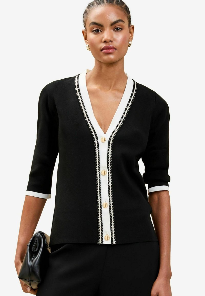 Svart cardigan med V-ringning, vit kant och dekorativa knappar. Har en figurnära design och trekvartslånga ärmar. Texturerat tyg.