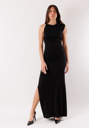 Donna in piedi con un abito nero senza maniche lungo fino a terra con uno spacco laterale, indossando tacchi neri, su uno sfondo chiaro uniforme.