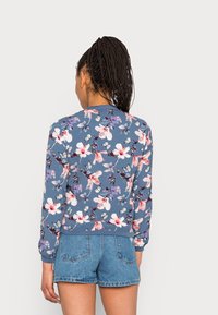 Felpa blu floreale con motivi di fiori rosa e bianchi, maniche lunghe e polsini a coste, abbinata a pantaloni corti di jeans.