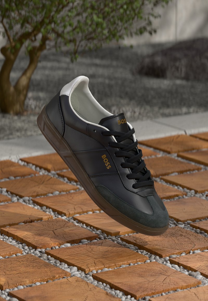 Adidași din piele neagră cu accente albe, design cu șireturi, vârf din suede de culoare verde oliu și logo-ul auriu "BOSS". Poziționați deasupra plăcilor de lemn cu pietriș.