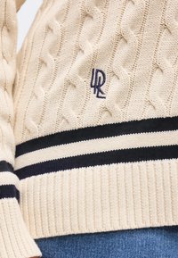 Κρεμ πουλόβερ με πλέξη καλώδιο και σκούρες μπλε οριζόντιες ρίγες, με κεντημένο λογότυπο "LRL" κοντά στο κάτω μέρος, φορεμένο με μπλε τζιν.