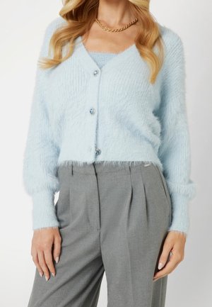 Cardigan bleu clair duveteux avec trois boutons décoratifs sur un haut assorti, associé à un pantalon plissé gris taille haute et un collier chaîne en or.