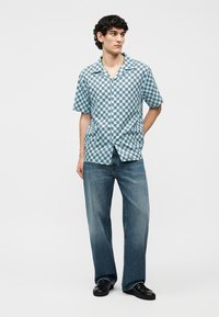 Young man with curly hair wearing a blue and white checkered short-sleeve shirt, loose blue jeans, and black shoes, standing with one hand behind.