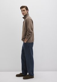 Brown fleece pullover with a high collar, paired with loose blue denim jeans and dark brown moccasin-style shoes.