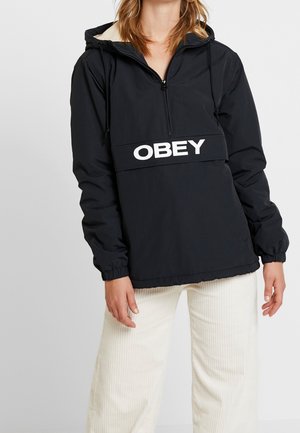 Person iført en sort hættetrøje med hvid "OBEY" tekst over brystet og lyse bukser.