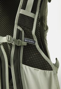 Zaino a rete verde con spallacci imbottiti e accessori regolabili. Logo Columbia visibile su tessuto beige con dettagli testurizzati.